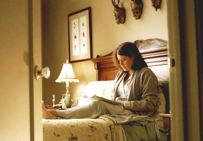 8 Dr. Melfi sitting on a bed reading a study under a lamp in a bedroom scene from the penultimate episode of The Sopranos.
