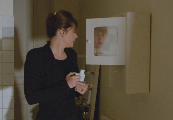 2 Lorraine Bracco in a black dress looking at a bathroom wall cabinet in Les Menteurs
